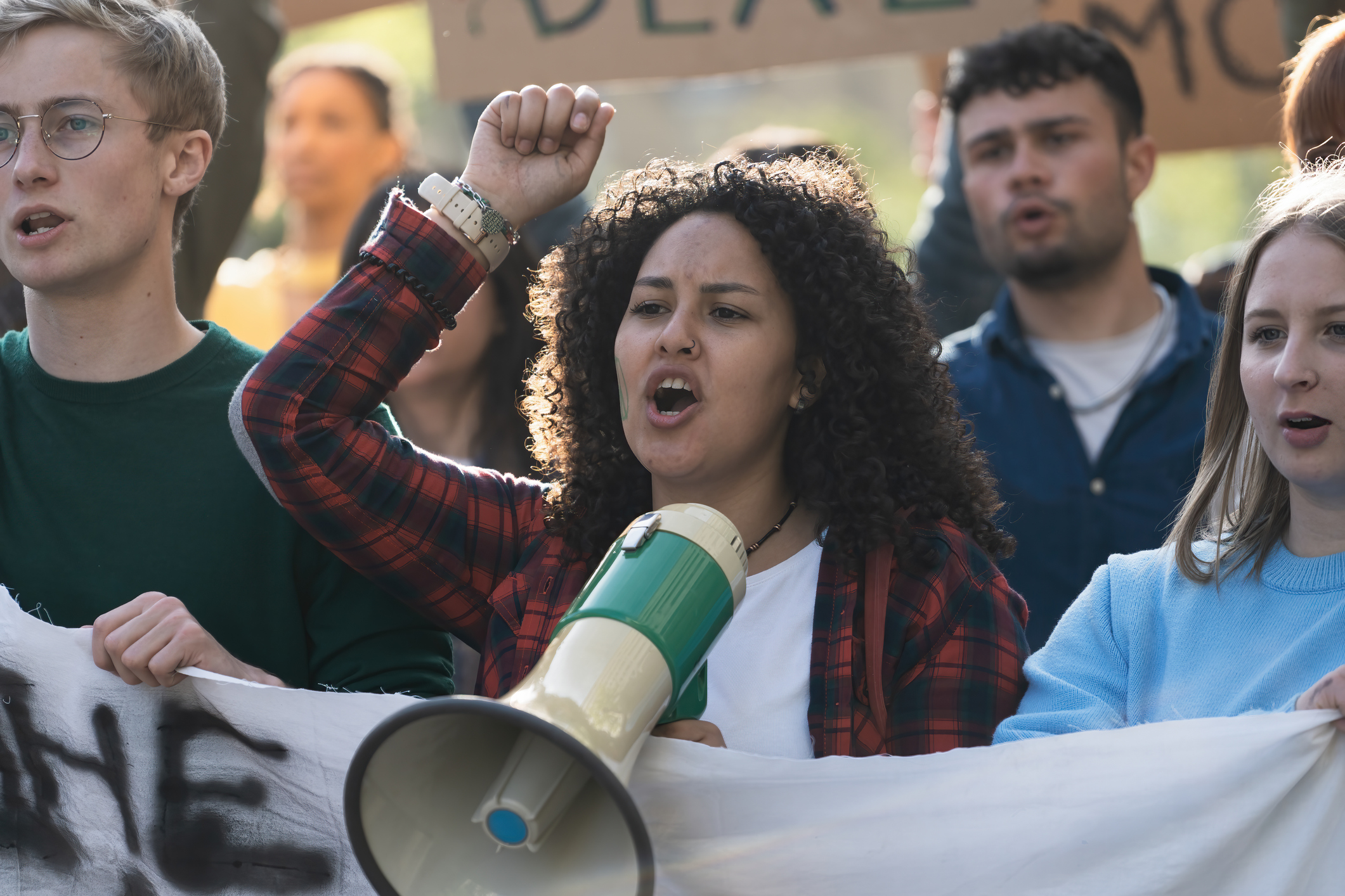 Close-up of a young woman with curly hair, leading a student protest using a megaphone. The group, holding an unidentifiable banner, passionately chants slogans.